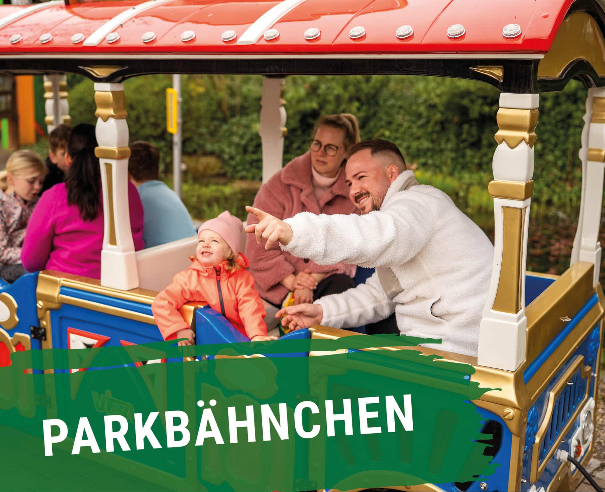 Familie mit Kleinkind fährt lachend im Parkbähnchen, Vater zeigt begeistert in die Ferne