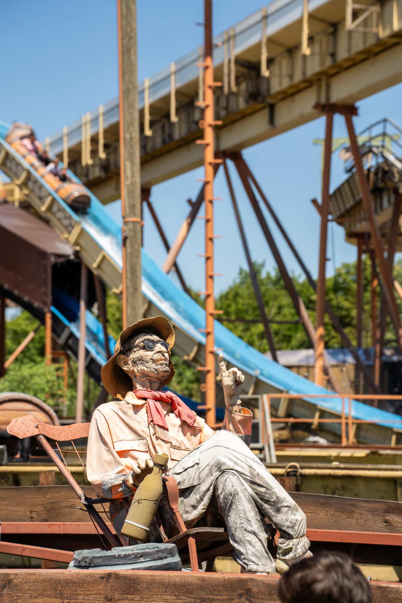 Animatronische Figur eines alten Mannes mit Hut und Sonnenbrille sitzt entspannt vor einer Wildwasserbahn im Freizeitpark