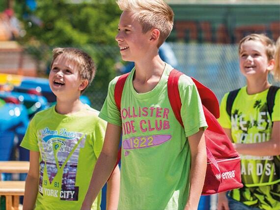 Drei Kinder im Freien, zwei tragen grüne T-Shirts und einer hat einen roten Rucksack.