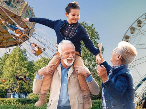 Ein Großvater trägt seinen Enkel auf den Schultern, während eine Großmutter lächelt. Im Hintergrund sind Fahrgeschäfte sichtbar.