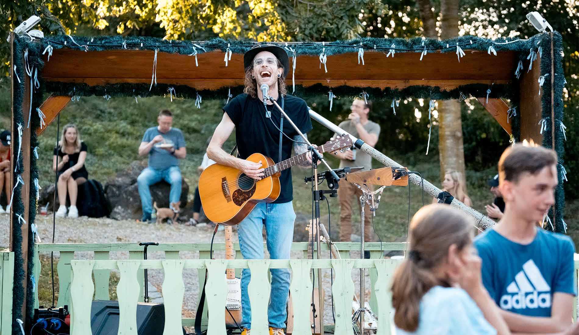 Ein Mann mit Hut spielt Gitarre und singt auf einer kleinen Bühne im Freien.