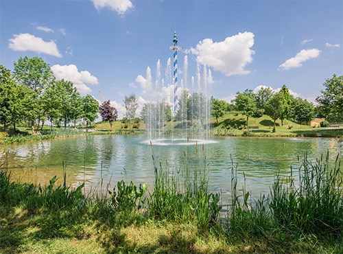 Ein See mit einer Wasserfontäne im Allgäu Skyline Park, umgeben von Bäumen und Gras.