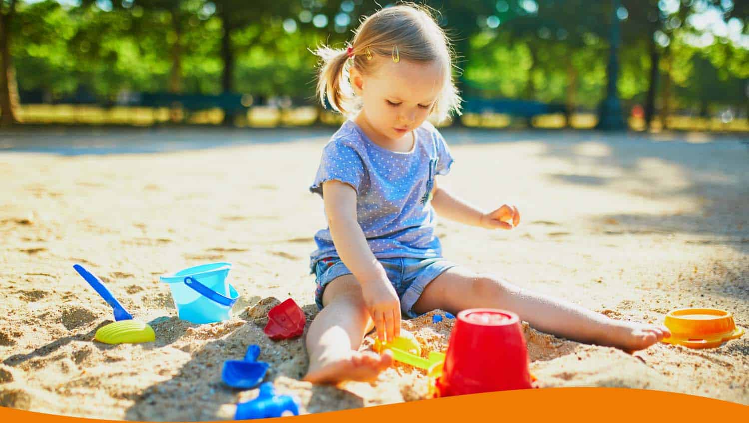 Ein kleines Kind sitzt im Sandkasten und spielt mit buntem Spielzeug, darunter Eimer und Förmchen.