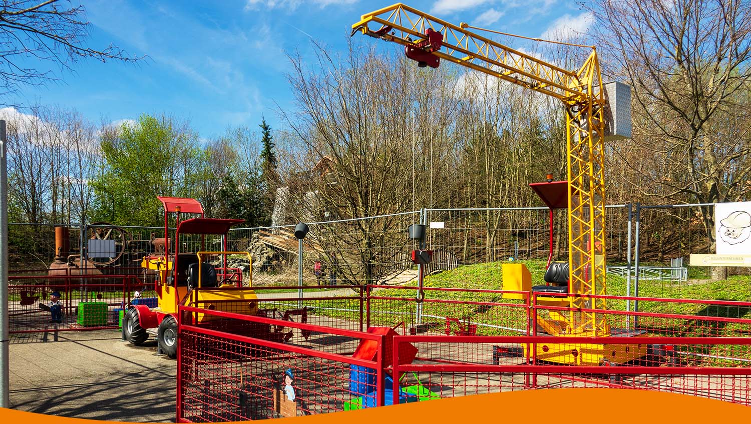 Gelber Kran mit Spielzeugfahrzeugen auf einem Spielplatz, umgeben von einem Zaun.