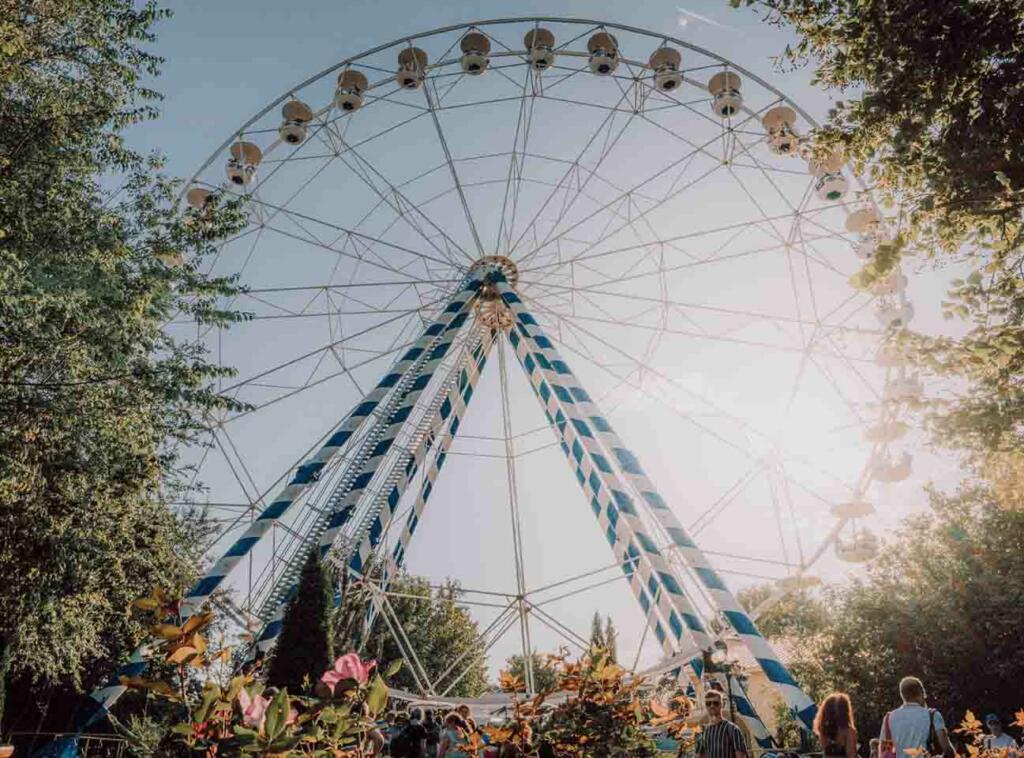 Riesenrad mit blauen und weißen Streifen, umgeben von Bäumen und Sonnenlicht.