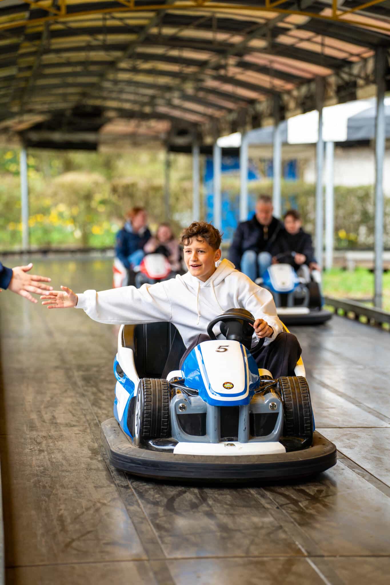 Junge fährt lachend in einem blauen Elektro-Bumper-Car auf einer überdachten Bahn, weitere Fahrer im Hintergrund