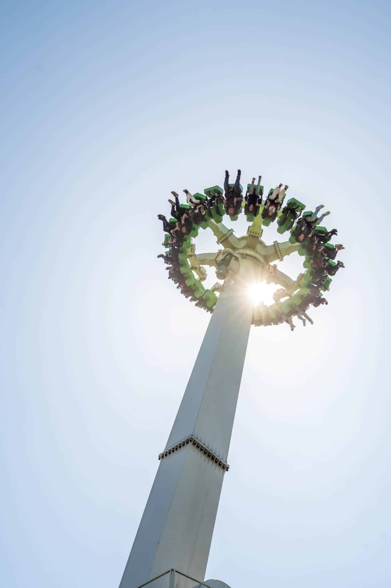 Fahrgäste in grünen Sitzen eines Freifall-Karussells drehen sich kopfüber auf einem hohen Turm vor blauem Himmel