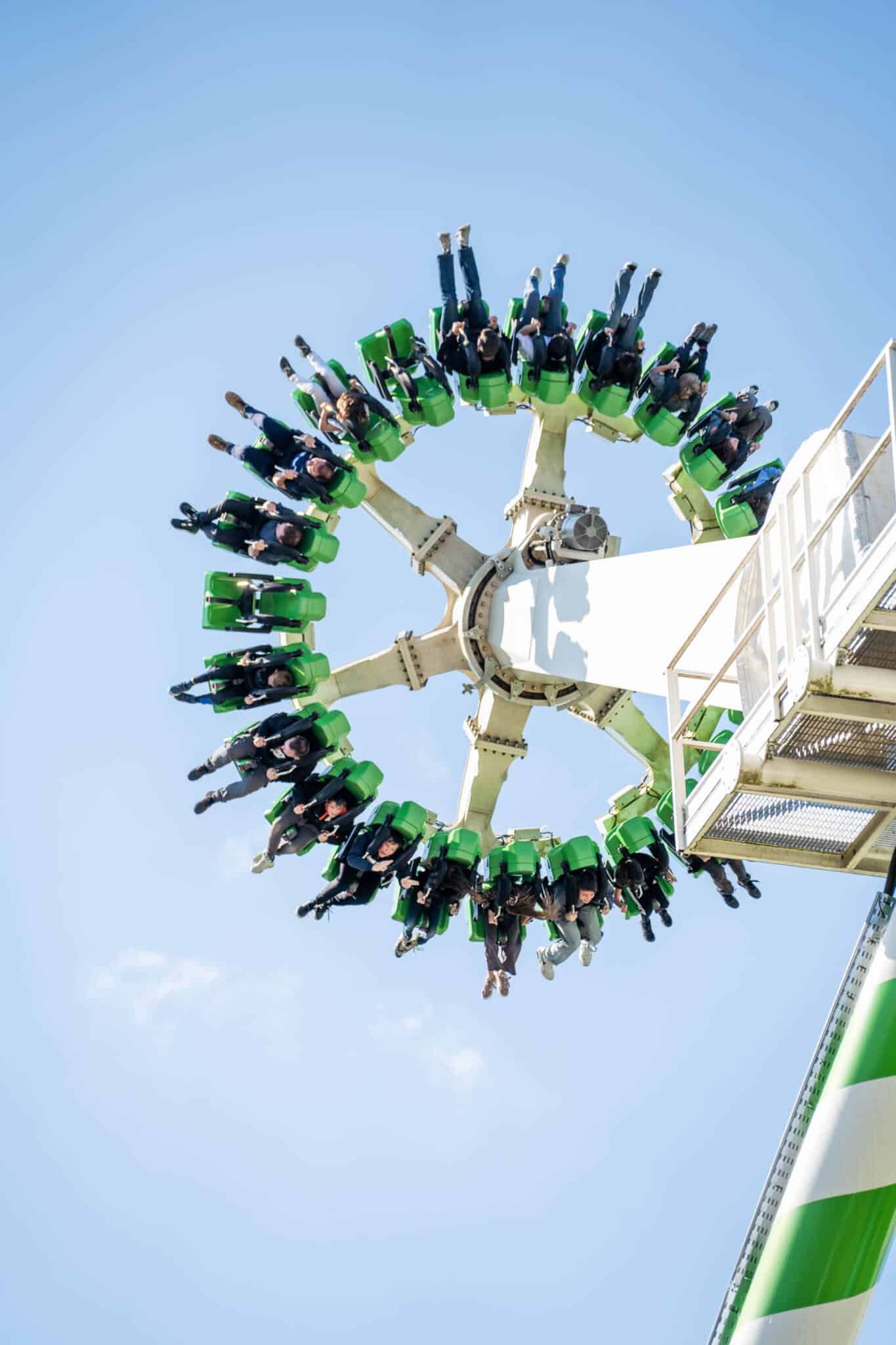 Fahrgäste in grünen Sitzen eines Freifall-Karussells drehen sich kopfüber auf einem hohen Turm vor blauem Himmel