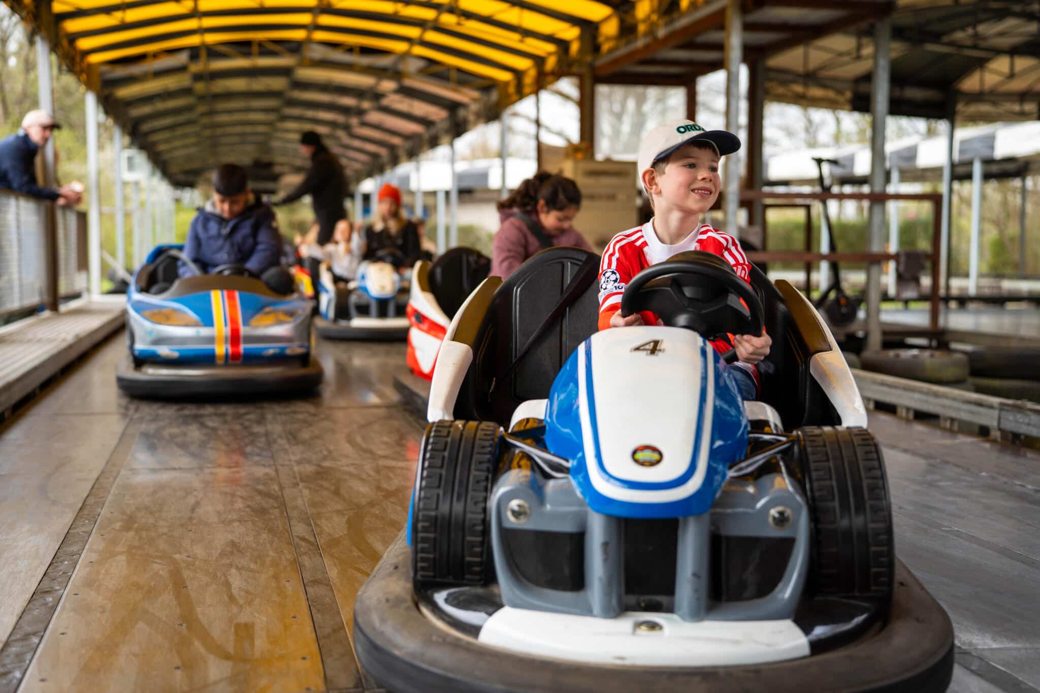 Kind fährt lächelnd ein Go-Kart auf einer überdachten Bahn, weitere Kinder und Erwachsene im Hintergrund