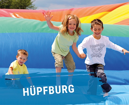 Drei Kinder springen auf einer bunten Hüpfburg mit Regenbogenfarben.