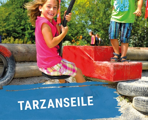 Ein Kind schwingt auf einer Tarzan-Seilbahn auf einem Spielplatz, während ein anderes Kind daneben steht.