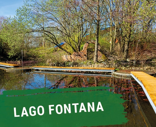 Wasserfläche Lago Fontana mit Holzstegen und Bäumen im Hintergrund.
