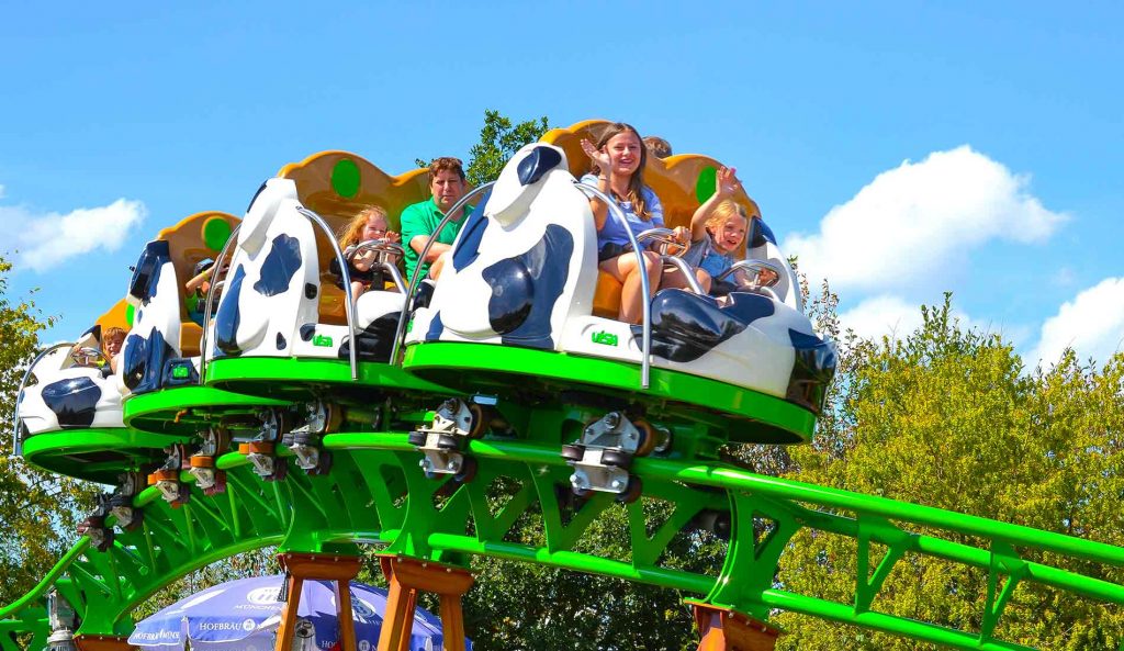 Achterbahn mit Kuhdesign, auf der mehrere Personen sitzen, im Freizeitpark unter blauem Himmel.